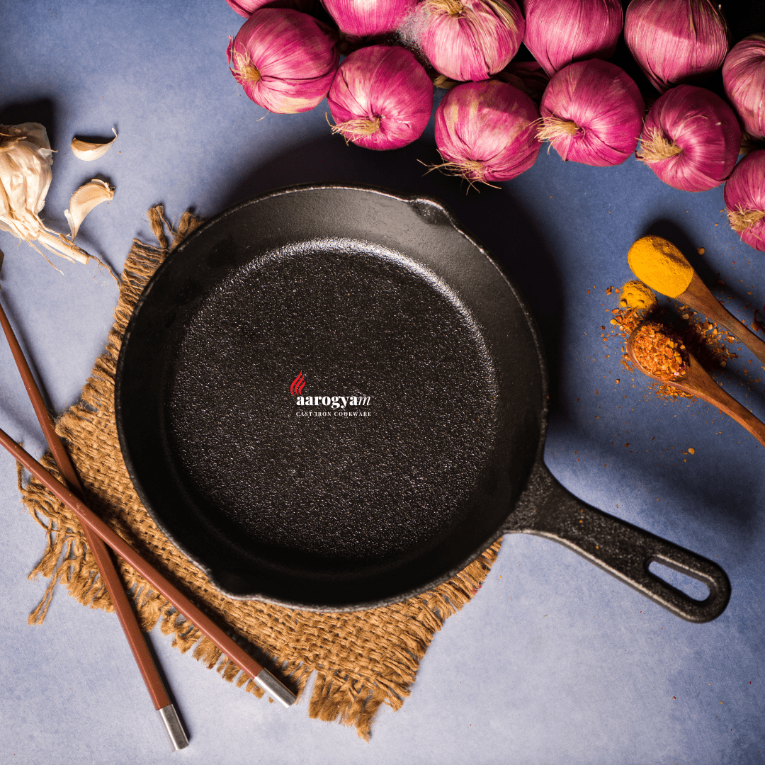 Black cast iron skillet with 'Aarogyan' branding on a blue surface with pink onions and spices.
