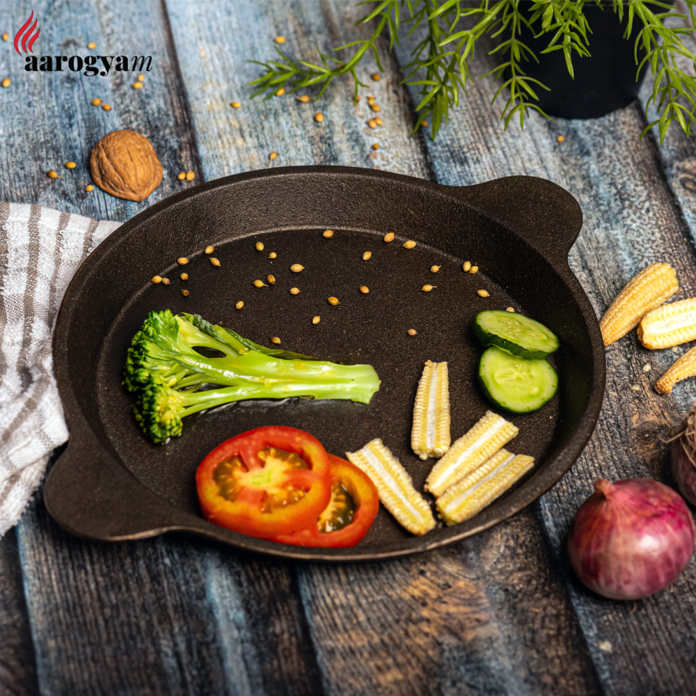 Black cast iron skillet with vegetables on a wooden surface, featuring the brand 'aarogya'.