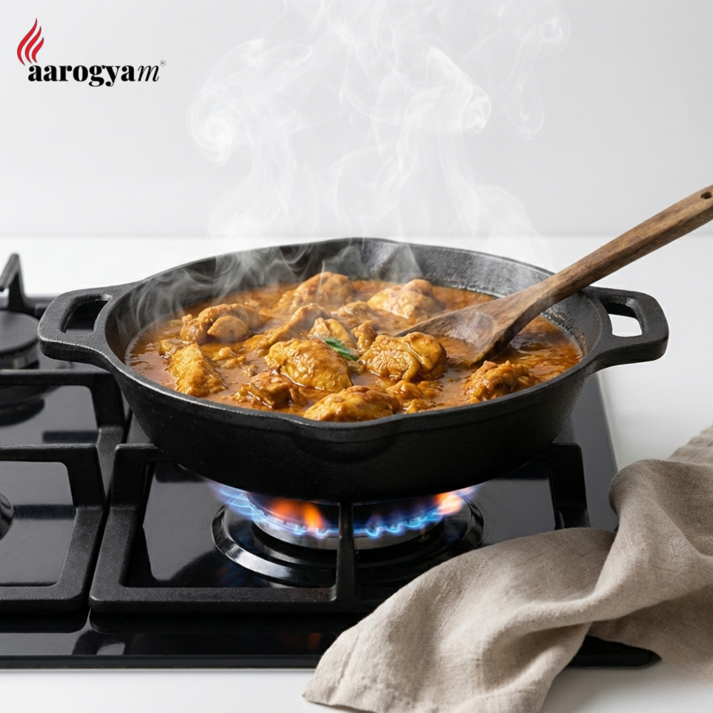 Stir-frying chicken curry in a black cast iron skillet on a gas stove with 'aarogya' branding.