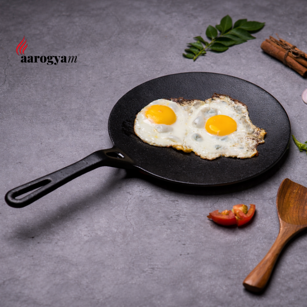 Fried eggs on a black cast iron pan with a wooden spatula and spices on a gray background, featuring the 'aarogyam' brand.