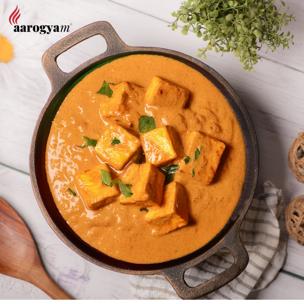 Dish of tofu in a red curry sauce with a wooden spoon and plant on a white surface, branded 'aarogya'.