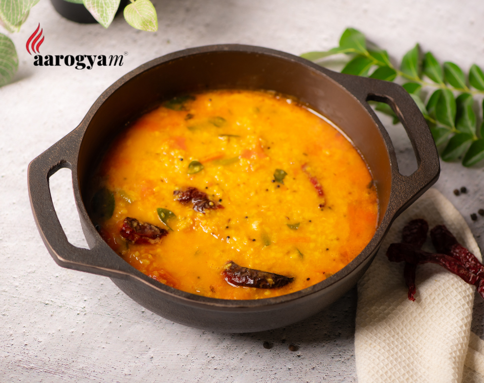 Yellow lentil soup in a black pot with a white napkin and green leaves on a light gray background, featuring the 'aarogyam' brand.