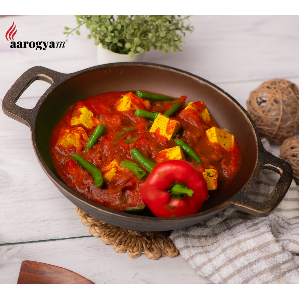 Dish of tofu and vegetables in a red sauce in a cast iron skillet on a light wooden surface with 'aarogya' branding.