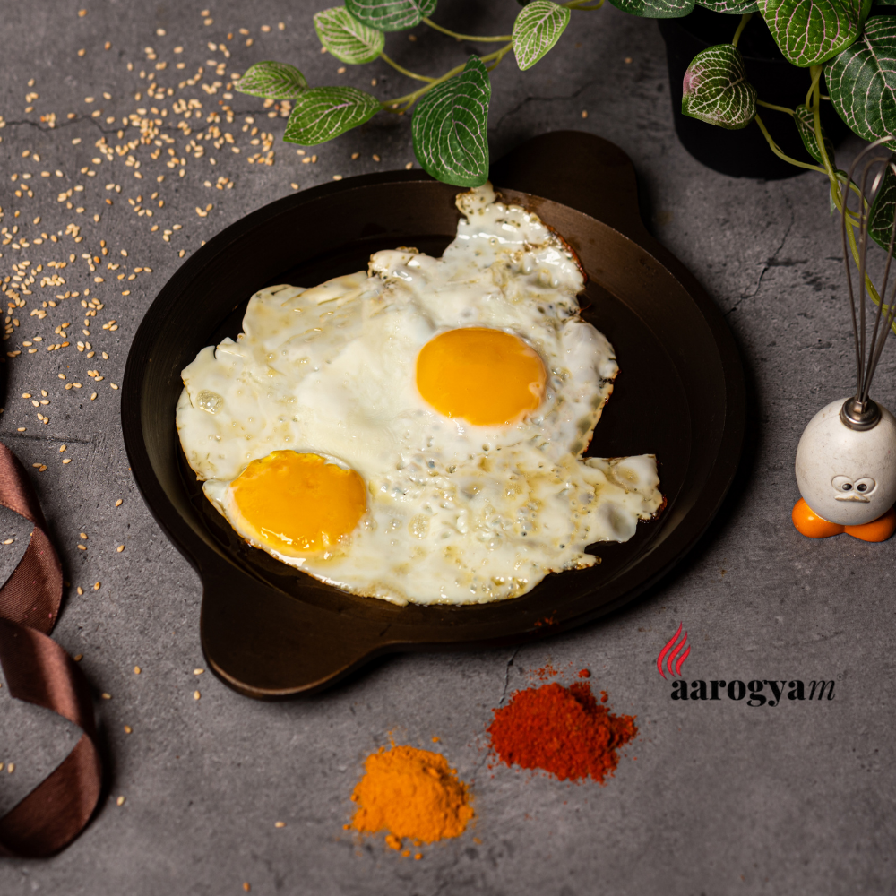 Two sunny-side-up eggs on a black plate with spices and a plant in the background, branded 'aarogyam'.