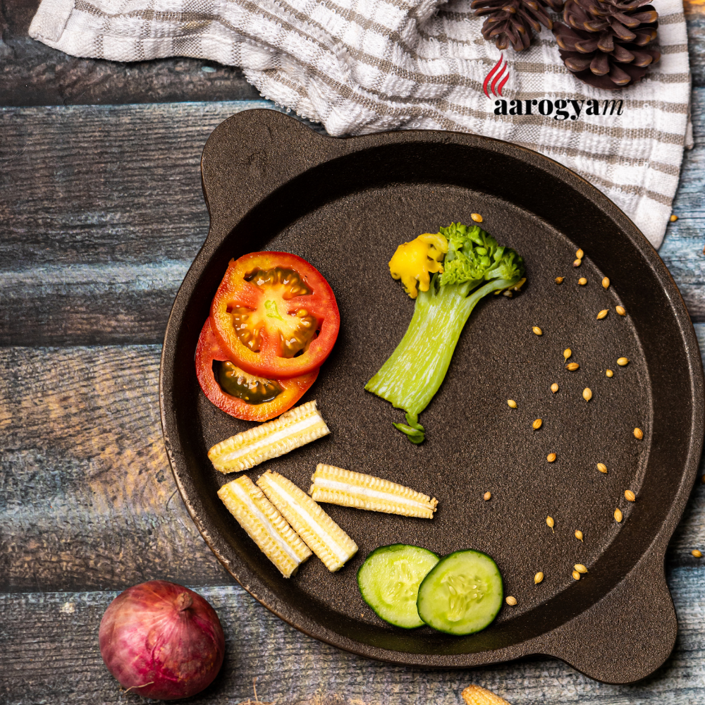Cast iron skillet with vegetables on a wooden surface, featuring the 'aarogya' brand.