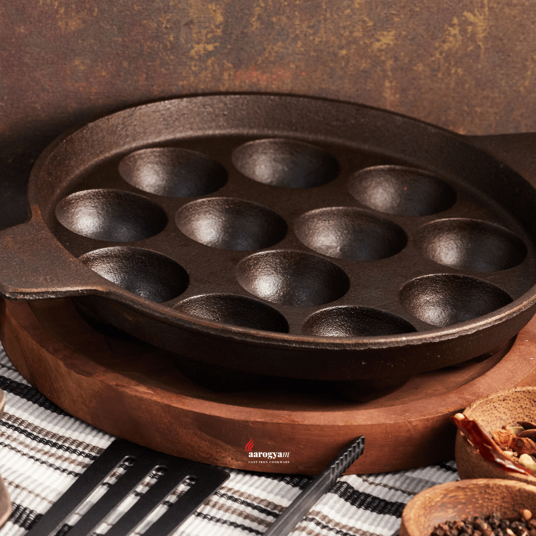 Cast iron cast iron appam patra pan with multiple small compartments on a wooden stand, brand 'Aarogyam' visible.