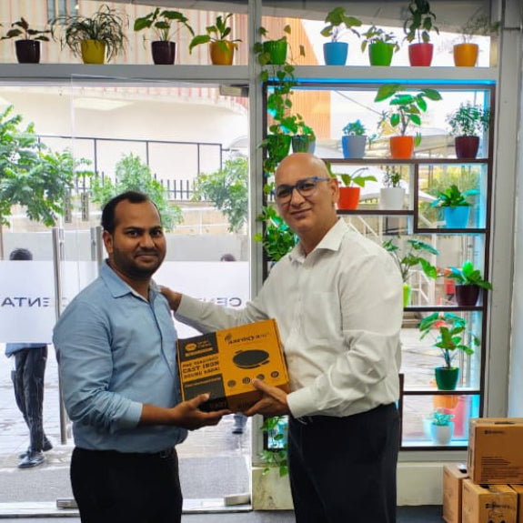 Two men holding an aarogyam cast iron cookware brand gift box of a cast iron kadhai as a diwali gift in an office with plants in the background